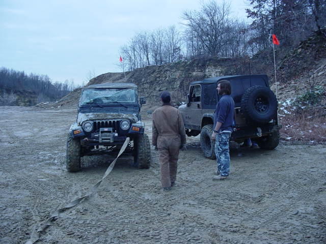 Two Jeep Wranglers, one with a broken windshield, are in a dirt lot with a tow strap connecting them