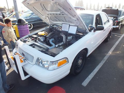 2003 Ford Crown Victoria - white Ford Crown Victoria with a front push bar and hood raised over a turbocharged engine, spectators standing nearby.