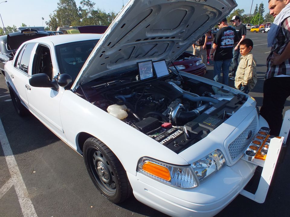 White Ford Crown Victoria with push bar, open hood and spectators at a car show