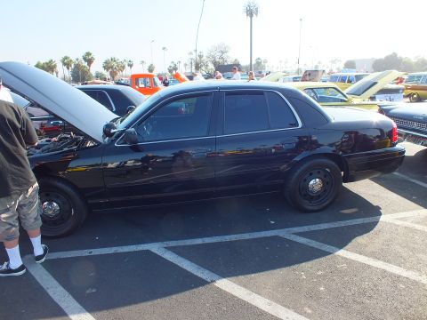 2003 Ford Crown Victoria - Side view of a black Ford Crown Victoria with its hood raised and black steel wheels, parked among other show cars.
