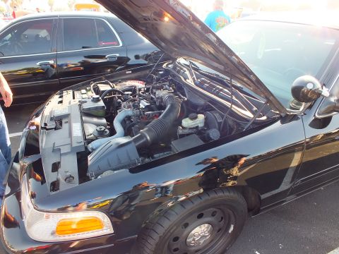 2003 Ford Crown Victoria - Front three-quarter view into the engine bay of a black Ford sedan with hood raised and intake plumbing visible at a show.