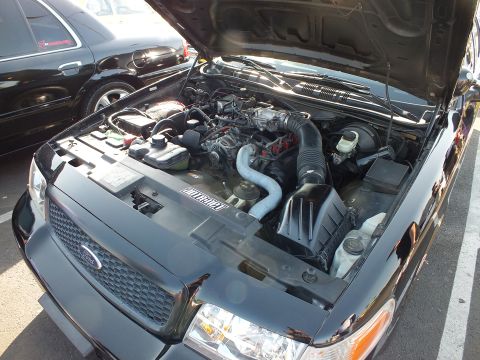 2003 Ford Crown Victoria - Engine bay of a dark Ford sedan with hood raised showing the air intake and plumbing, another dark sedan parked alongside.