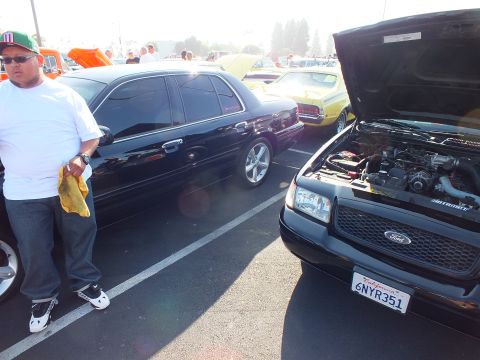 2003 Ford Crown Victoria - man in a white shirt stands by a black Ford with hood up and plate 6NYR351 next to another dark sedan at an outdoor show.
