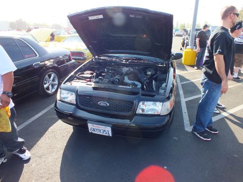 2003 Ford Crown Victoria - black Ford with raised hood and California plate 6NYR351, with spectators standing beside it at a sunlit car show.