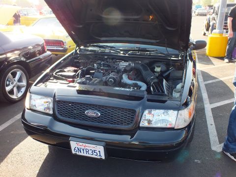 2003 Ford Crown Victoria - Front view of a black Ford with mesh grille, raised hood over the engine bay, and California plate 6NYR351 at a car show.