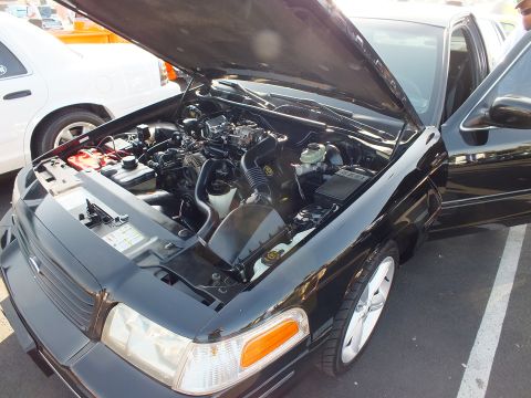 2003 Ford Crown Victoria - Front three-quarter view of a black Ford sedan with hood raised showing the engine, with a polished chrome wheel visible.