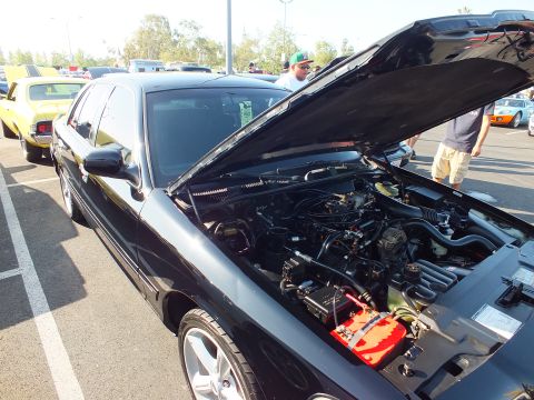 2003 Ford Crown Victoria - Front-side view of a black Ford sedan with hood raised showing the engine and a red battery box, parked beside a yellow classic car.