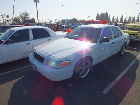 2003 Ford Crown Victoria - white Ford Crown Victoria sedan sits in a sunlit parking lot car show with other vehicles and palm trees in the background.