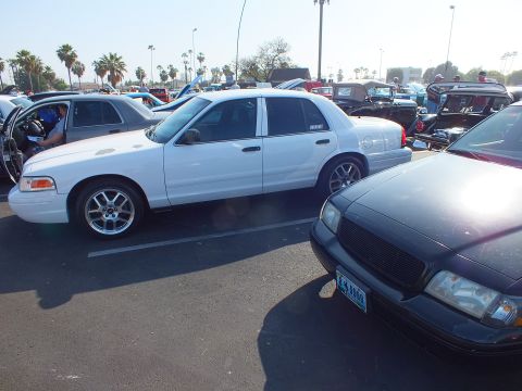 2003 Ford Crown Victoria - white Ford Crown Victoria on chrome wheels parked beside a dark gray sedan with a blacked-out grille at an outdoor car show.