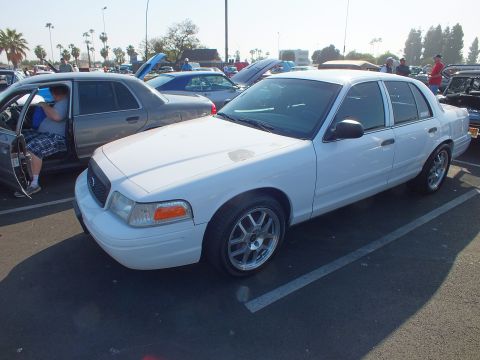 2003 Ford Crown Victoria - white Ford Crown Victoria parked at a car show as a man climbs into an adjacent gray sedan under a bright sky.