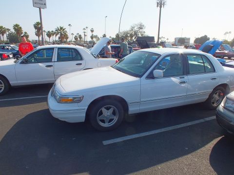 2003 Ford Crown Victoria - white full-size sedan on five-spoke alloy wheels parked beside another white sedan with its hood up at a car show.