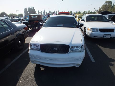 2003 Ford Crown Victoria - white Ford sedan photographed head-on, its Ford grille emblem visible, parked among other cars at an outdoor show.