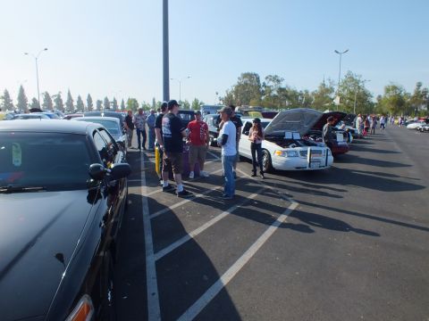2003 Ford Crown Victoria - Spectators gather around parked cars with raised hoods across a wide sunny parking lot at a car show.
