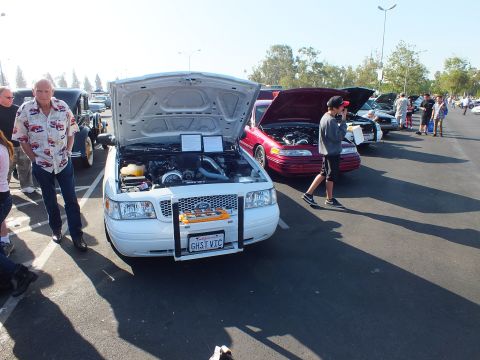 2003 Ford Crown Victoria - white sedan with raised hood and personalized plate GHST VIC is parked beside a red car as people browse the show.