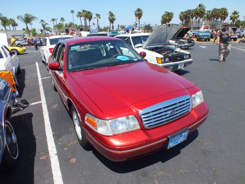 2003 Ford Crown Victoria - red Ford sedan with a chrome grille sits in a sunny lot lined with palm trees and other show cars.