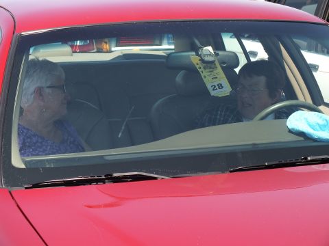 2003 Ford Crown Victoria - elderly couple sits inside a red car, seen through the windshield, with a number 28 placard hanging from the mirror.