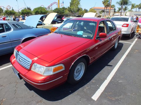 2003 Ford Crown Victoria - red Ford sedan with a chrome grille is parked in a show lot, a roller coaster visible beyond the palm trees.