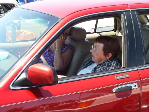 2003 Ford Crown Victoria - Two people sit inside a red sedan, viewed through the open driver-side window with the side mirror in the foreground.