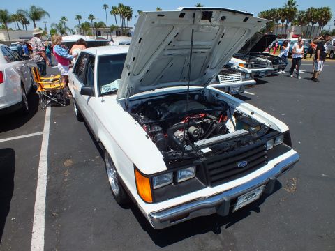 2003 Ford Crown Victoria - white Ford sedan sits with its hood raised revealing the engine bay, surrounded by other cars and spectators.