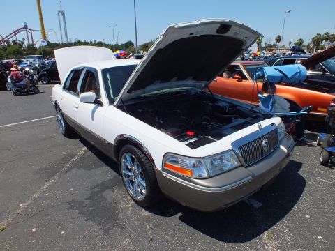 2003 Ford Crown Victoria - two-tone white and tan Mercury sedan displays its engine with hood up, a roller coaster looming in the background.