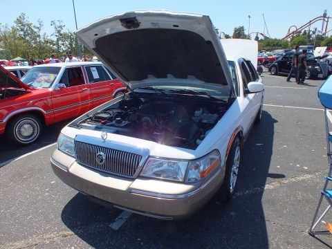2003 Ford Crown Victoria - two-tone Mercury sedan displays its engine bay with hood up, a roller coaster and show tents in the background.