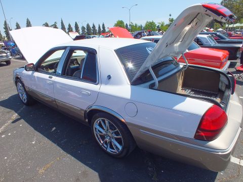 2003 Ford Crown Victoria - two-tone white and tan sedan with chrome wheels sits with both hood and trunk open at a crowded outdoor car show.