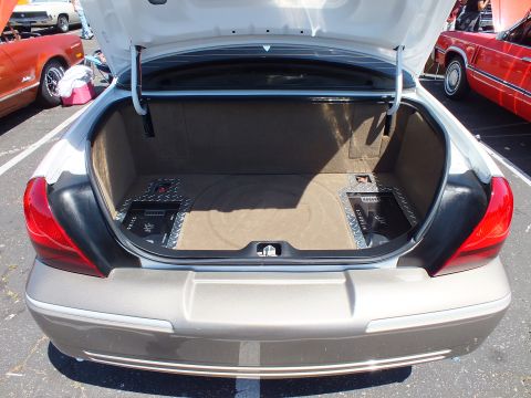 2003 Ford Crown Victoria - carpeted trunk of a sedan houses two car audio amplifiers, lid raised at a car show with red cars nearby.