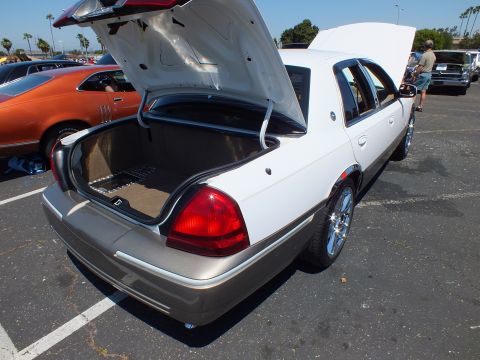 2003 Ford Crown Victoria - two-tone white and tan sedan with chrome wheels viewed from the rear, hood and trunk open at a sunny car show.