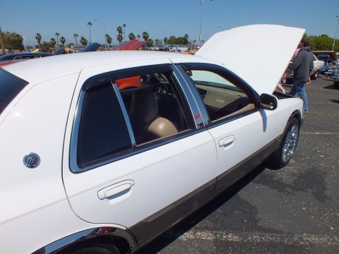 2003 Ford Crown Victoria - White four-door full-size sedan parked at a car show with its hood open, showing tan interior and chrome wheels.