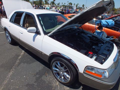 2003 Ford Crown Victoria - White four-door full-size sedan with chrome wheels and its hood raised, parked among other cars at an outdoor show.