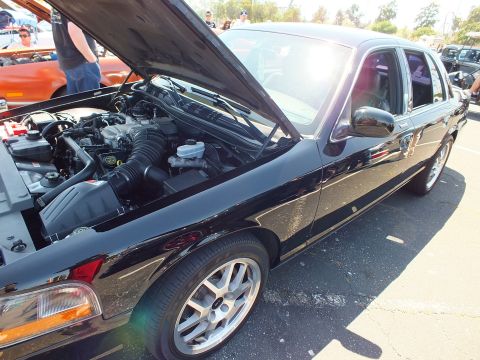 2003 Ford Crown Victoria - Black four-door sedan parked at a car show with its hood raised, showing the engine bay and silver alloy wheels.