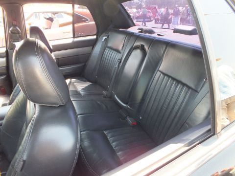 2003 Ford Crown Victoria - Rear bench seat of a sedan upholstered in black leather, viewed through the open rear door at an outdoor event.