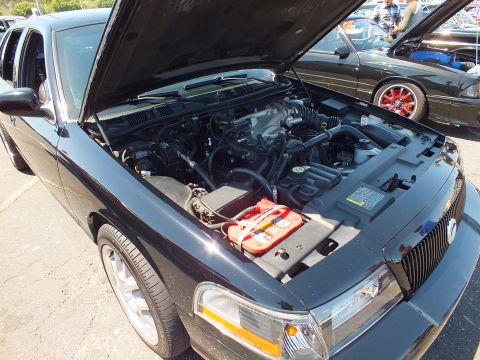 2003 Ford Crown Victoria - Open engine bay of a black sedan revealing the engine, a red battery cover, and chrome wheels, at a car show.
