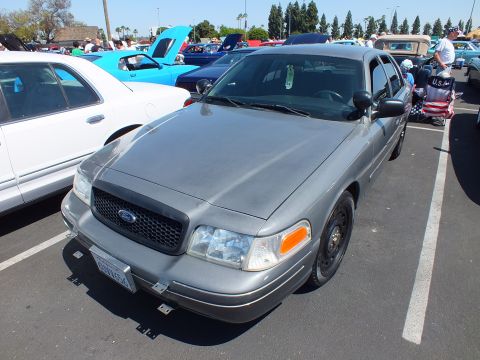 2003 Ford Crown Victoria - gray Ford sedan with the Ford emblem on its grille parked at an outdoor car show among colorful display cars.
