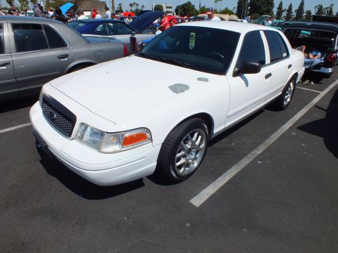 2003 Ford Crown Victoria - white Ford sedan with chrome multi-spoke wheels parked among other vehicles at a sunny outdoor car meet.