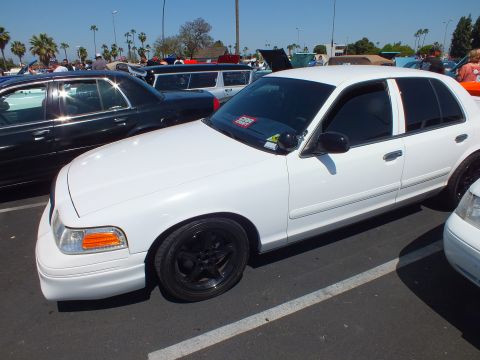 2003 Ford Crown Victoria - white Ford sedan with black steel-look wheels parked in a lot at a sunny outdoor car show.