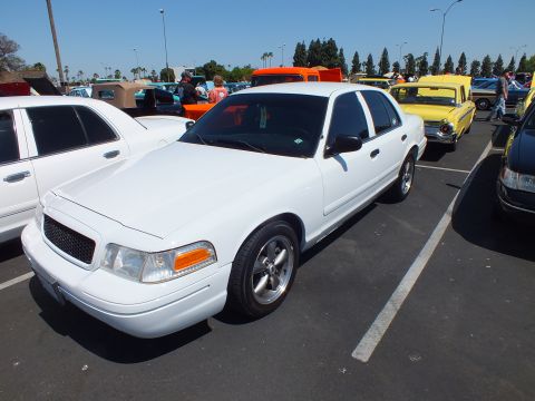 2003 Ford Crown Victoria - white Ford sedan with machined-face wheels parked beside a yellow vintage car at an outdoor car meet.