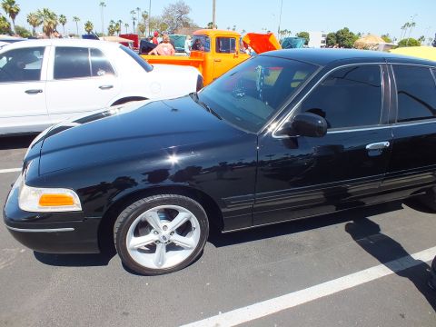 2003 Ford Crown Victoria - side profile of a black Ford sedan with chrome wheels parked near an orange pickup at an outdoor car show.
