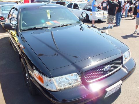2003 Ford Crown Victoria - black Ford sedan with a sun-lit chrome grille and Ford emblem parked among crowds at an outdoor car show.
