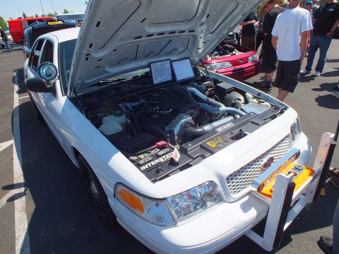 2003 Ford Crown Victoria - white Ford sedan with hood raised, a laptop on the engine and a flame-painted front bumper on display at a car show.