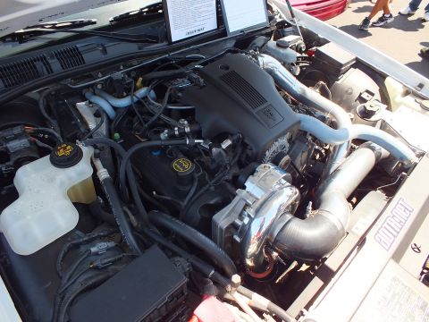 2003 Ford Crown Victoria - Engine bay of a white sedan showing a modified Ford V8 with chrome intercooler piping and a supercharger setup at an outdoor car show.