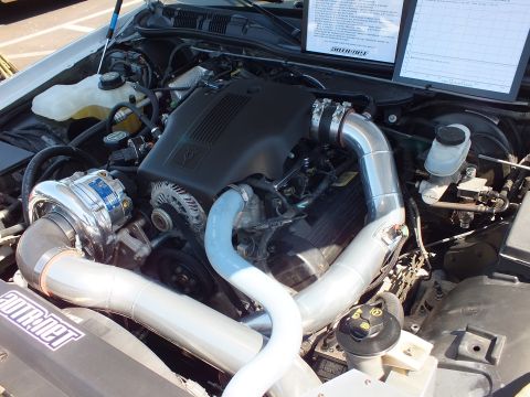 2003 Ford Crown Victoria - Open engine bay of a white sedan showing a supercharged Ford V8 with a polished intercooler, large piping and a printed display card.