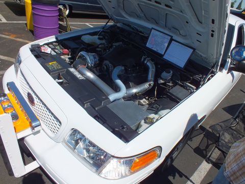 2003 Ford Crown Victoria - Front of a white vehicle with its hood open, fitted with a push bar and a modified engine with intercooler piping, beside a display card.