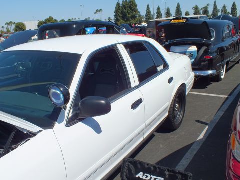 2003 Ford Crown Victoria - white Ford Crown Victoria sedan with a pillar spotlight and drag-style rear wheel parked in a lot next to vintage cars at a car show.