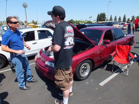 2003 Ford Crown Victoria - Two men converse next to a dark red sedan with its hood up and a red folding chair at a sunny outdoor car show.