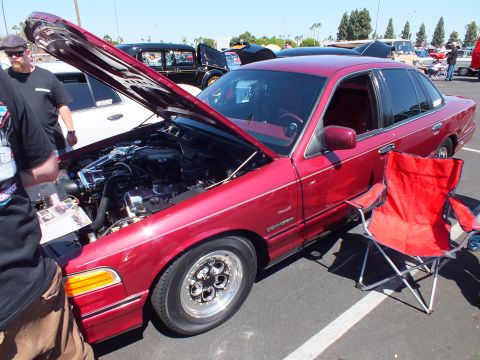 2003 Ford Crown Victoria - dark red Ford sedan with its hood open showing the engine bay, riding on polished slotted wheels, displayed at a car show.