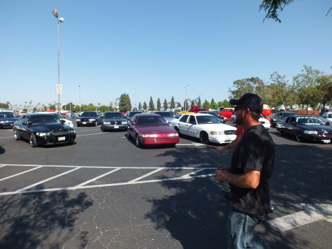 Man in a tie-dye shirt and cap standing in a parking lot of parked sedans