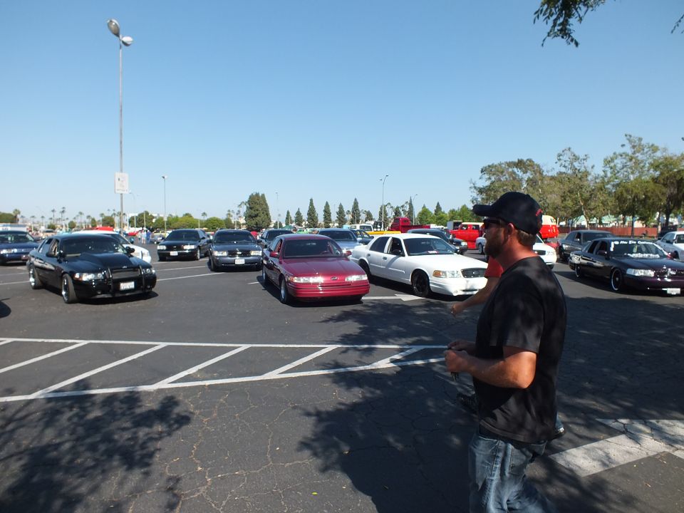 Man in a tie-dye shirt and cap standing in a parking lot of parked sedans