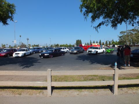 Parked cars, a green motorcycle and people gathered in a sunny lot
