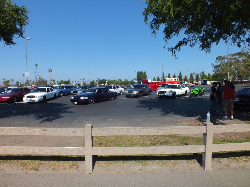 Parked cars, a green motorcycle and people gathered in a sunny lot